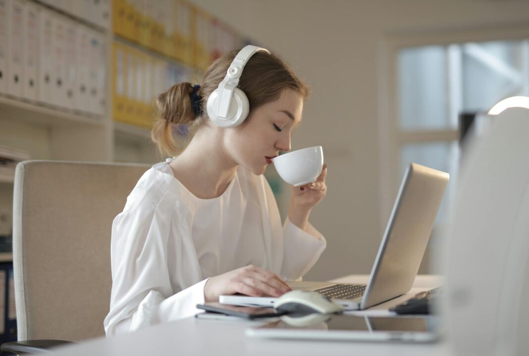 Woman wearing headphones and sipping coffee while working on a laptop at a desk.