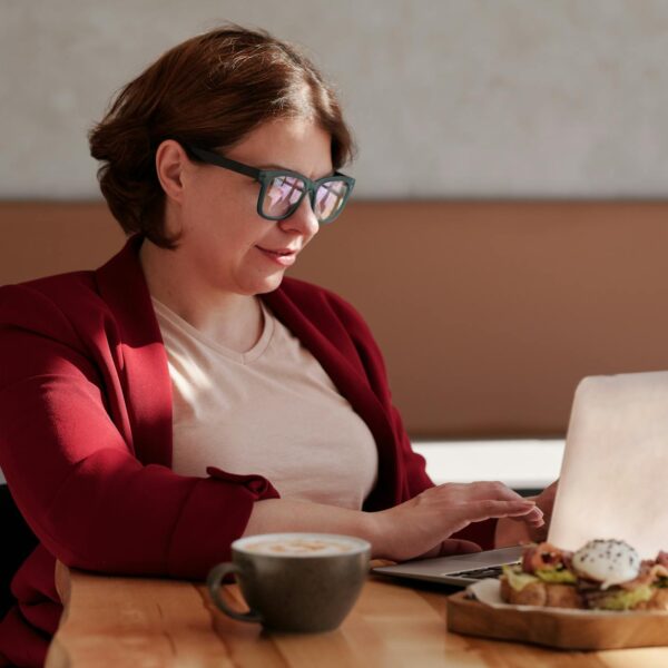Woman in a red blazer working on laptop at café table with coffee and snack.